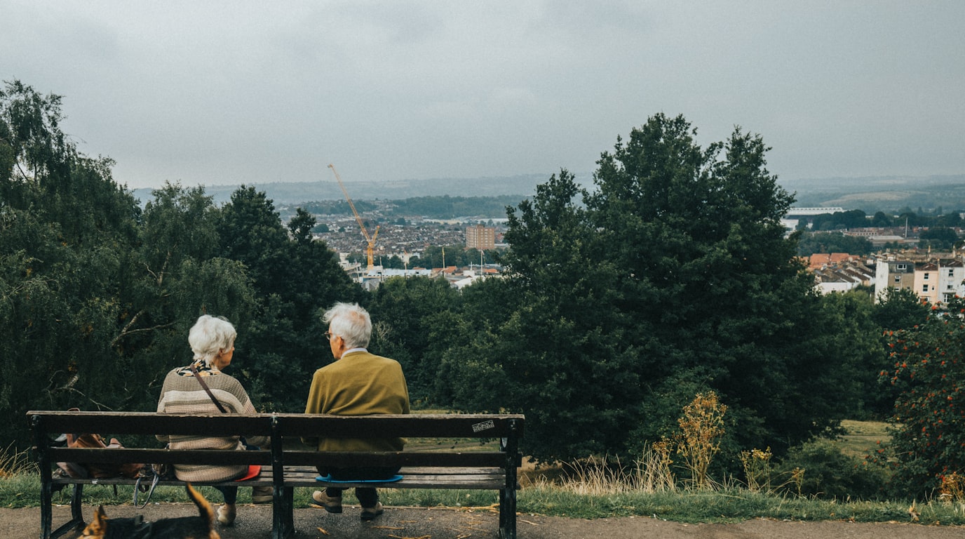 An older couple sitting together on a bench looking out over their town