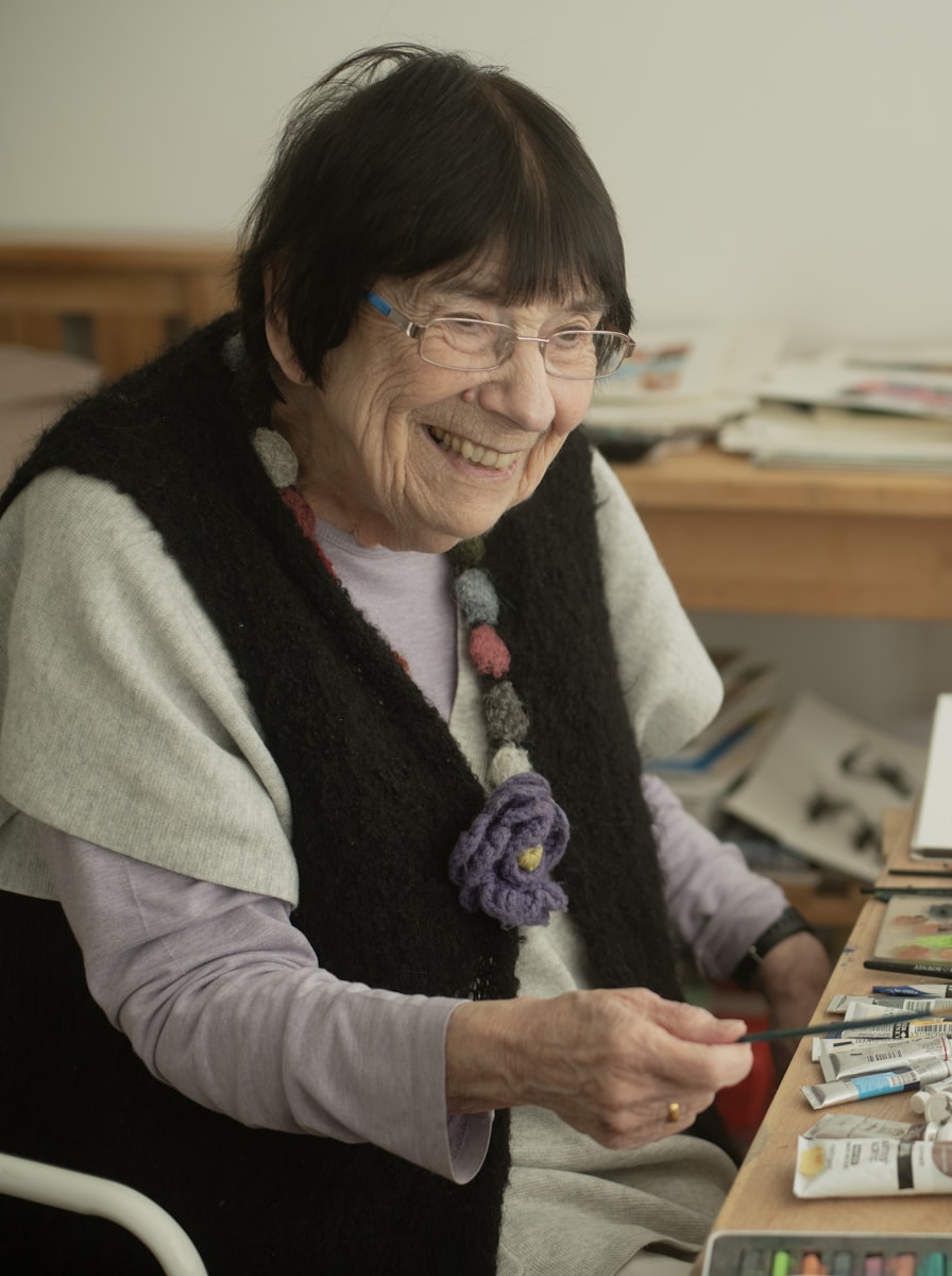 An older woman smiling at her worktable, surrounded by her paints